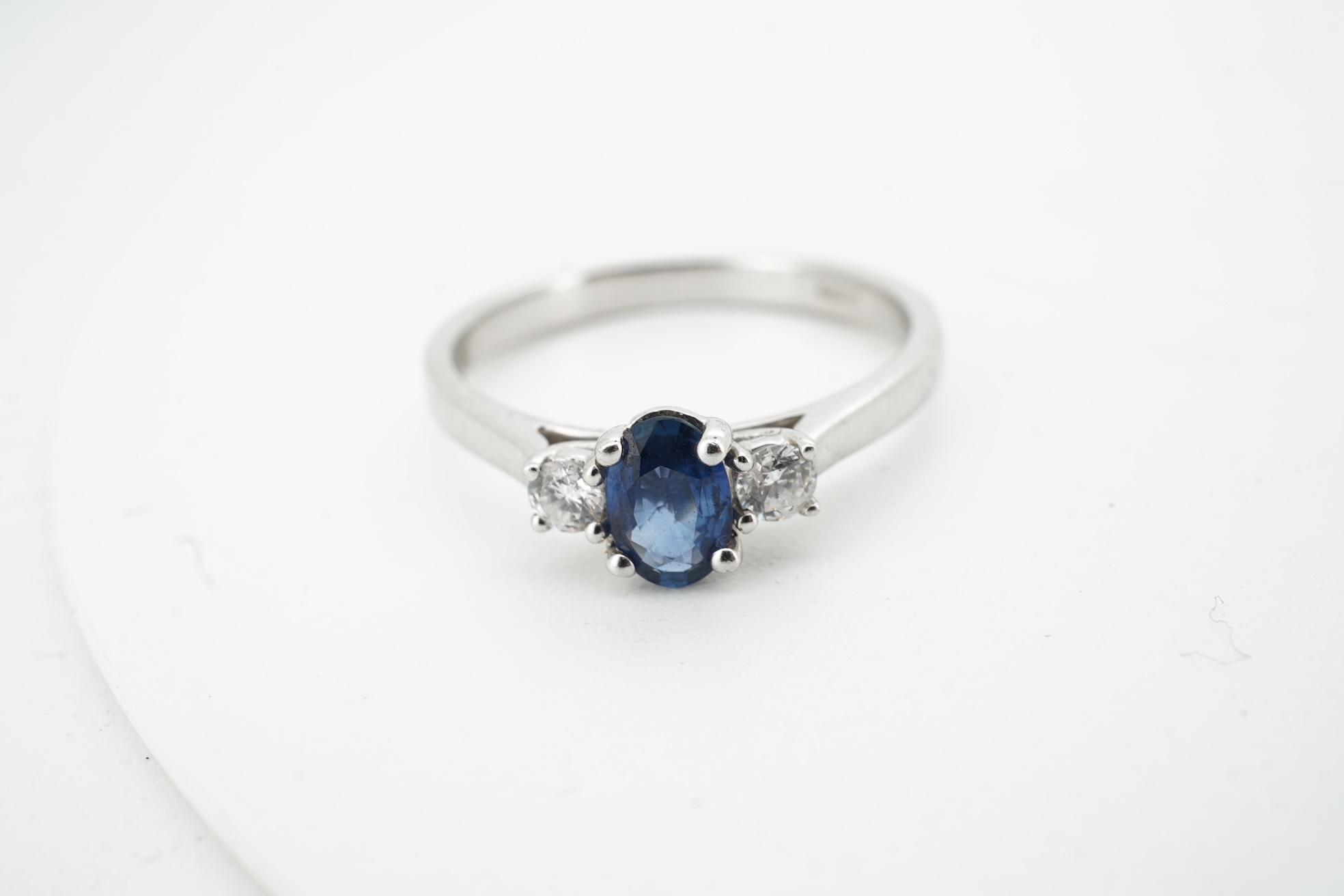 A sapphire and diamond three-stone ring, claw-set with an oval sapphire weighing approximately 0.35 carats, between shoulders set with brilliant-cut diamonds totalling approximately 0.12 carats, mounted in 9ct white gold